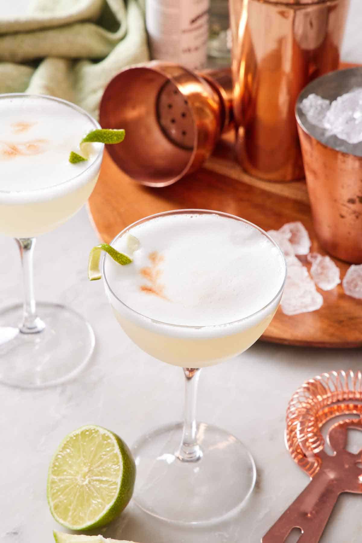 Two glasses of pisco sour cocktails with a lime garnish. Shaker in the background along with ice and some cut limes.