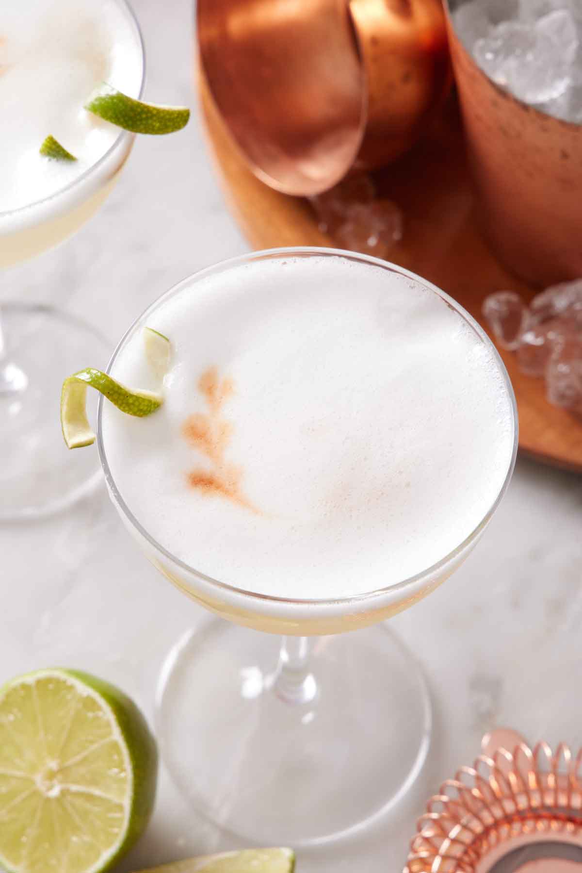 A slightly overhead view of a glass of pisco sour with a lime garnish. A shaker of ice off to the side and some cut limes and a second cocktail.