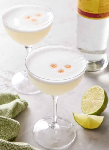 Two coupe glasses of pisco sour with cut limes and a bottle of pisco in the background.