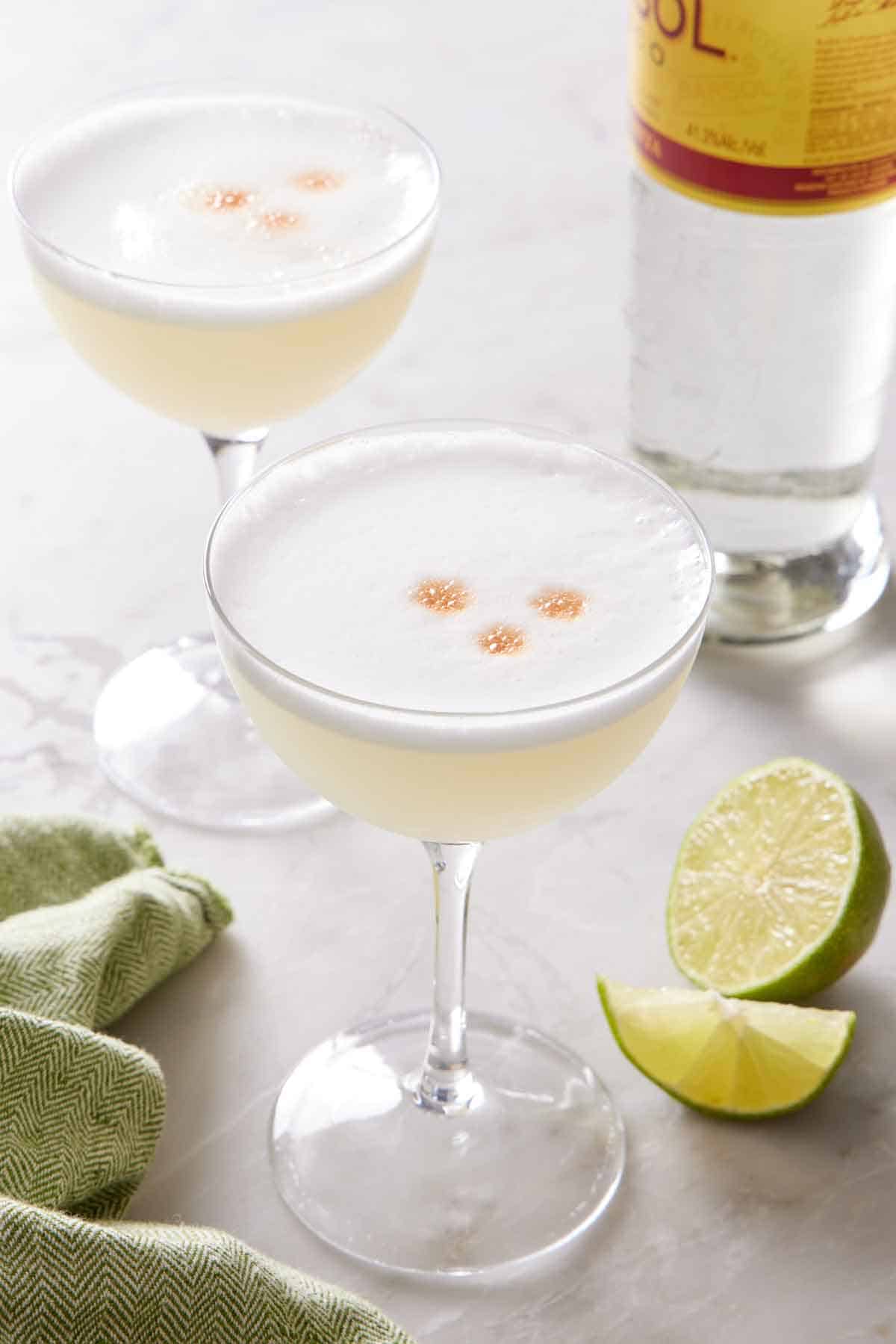 Two coupe glasses of pisco sour with cut limes and a bottle of pisco in the background.