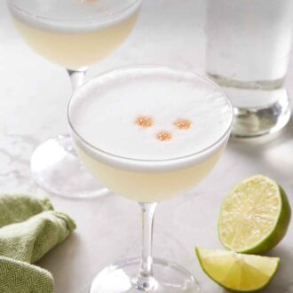 Pinterest graphic of two coupe glasses of pisco sour with cut limes and a bottle of pisco in the background.