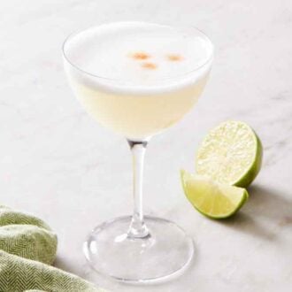 A coupe glass of a pisco sour cocktail with cut limes and a green napkin on the side.