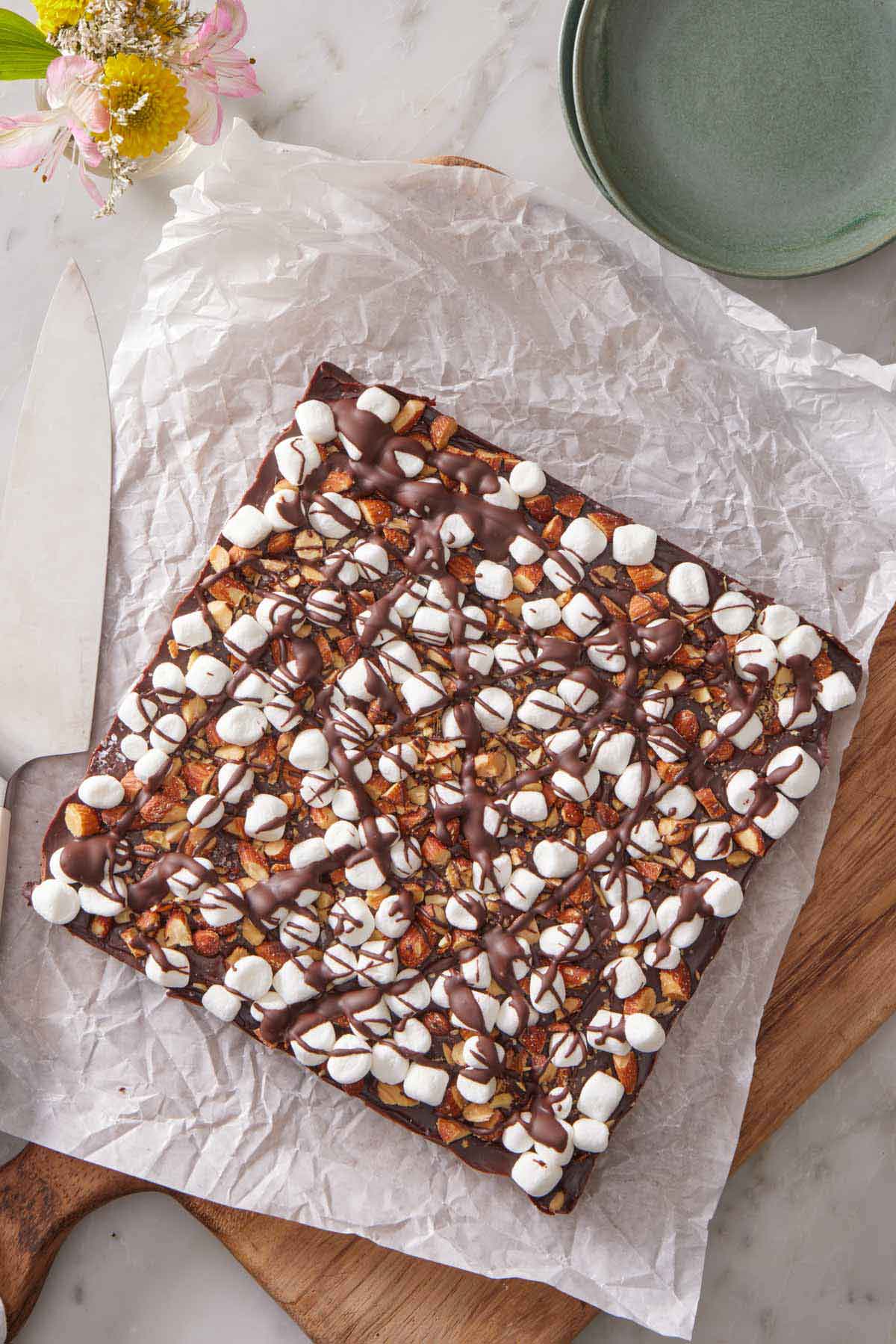 Overhead view of an uncut slab of rocky road bars on a parchment lined serving board. A knife on the side with a stack of plates.