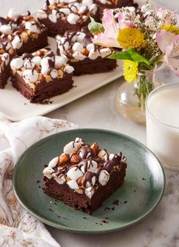 A plate with a rocky road bar with more on a platter in the background with a vase of flowers and glass of milk.