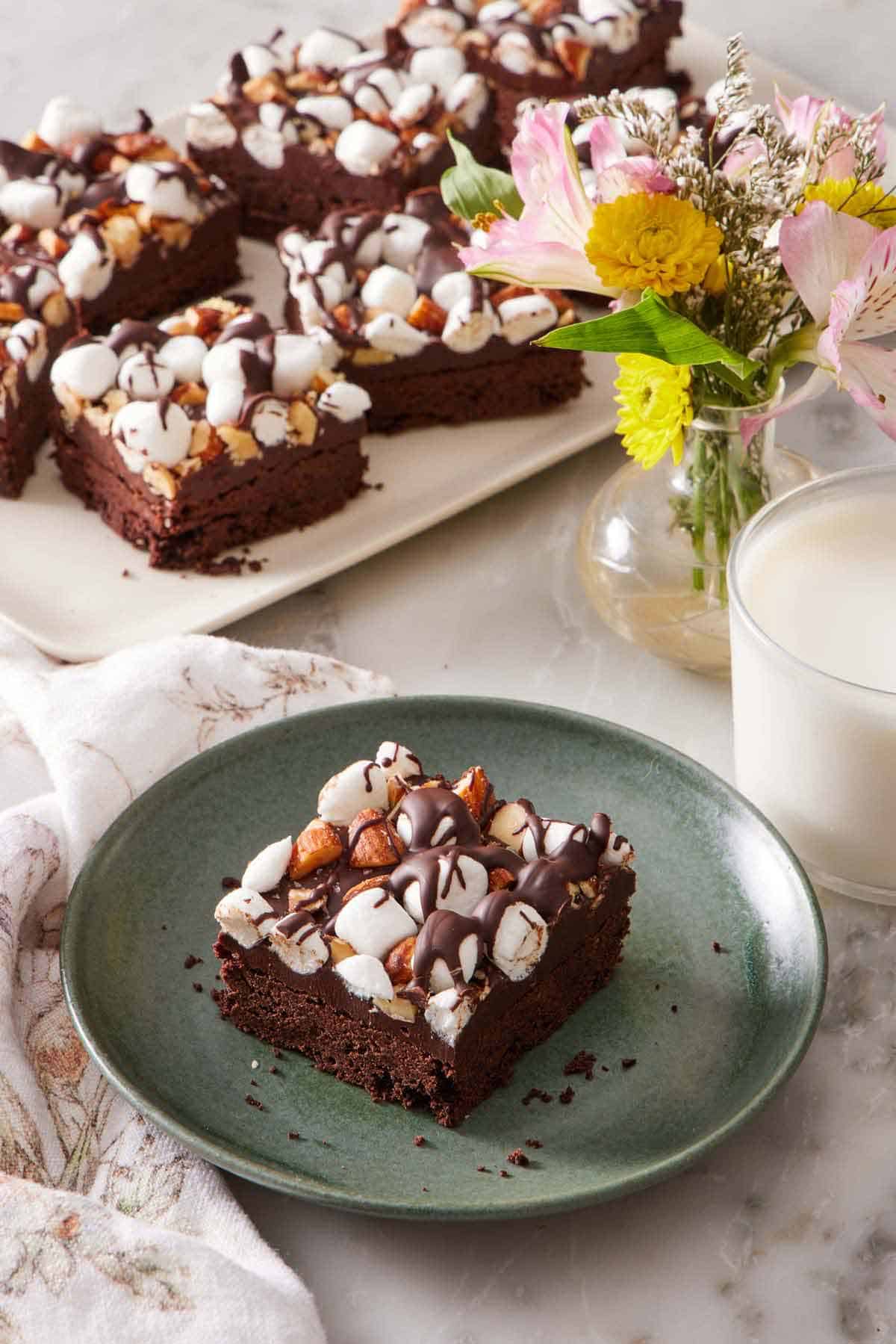 A plate with a rocky road bar with more on a platter in the background with a vase of flowers and glass of milk.