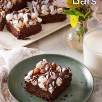 Pinterest graphic of a rocky road bar on a plate with a platter in the background with a vase of flowers and glass of milk.