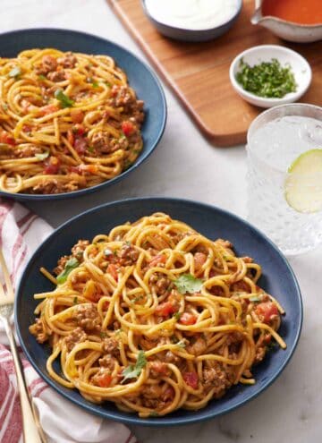 Two bowls of taco spaghetti with a drink, bowl of chopped cilantro, and two sauces in the background.