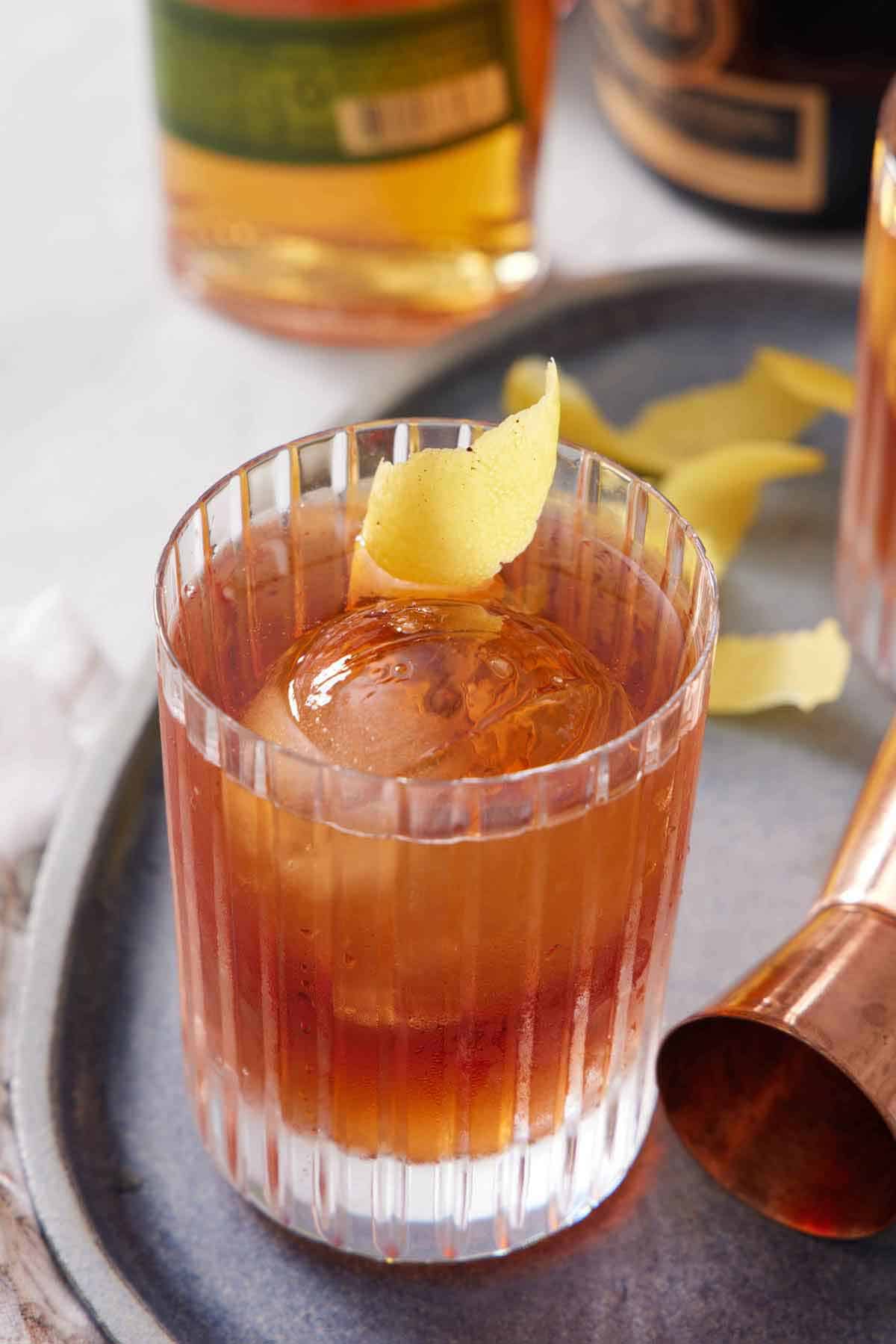 A glass of Vieux Carré with a round ice cube and a lemon peel garnish. A jigger on the side and more peels in the background.