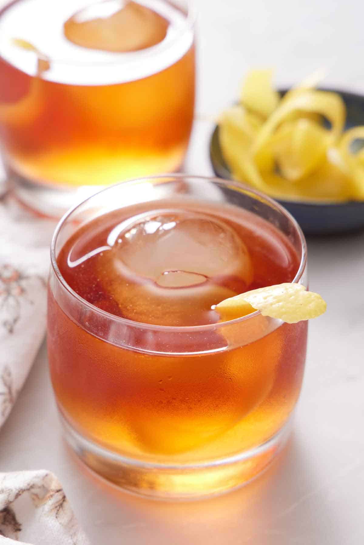 A glass of Vieux Carré with one ice cube with a lemon peel garnish. Another drink in the back along with more lemon peels.
