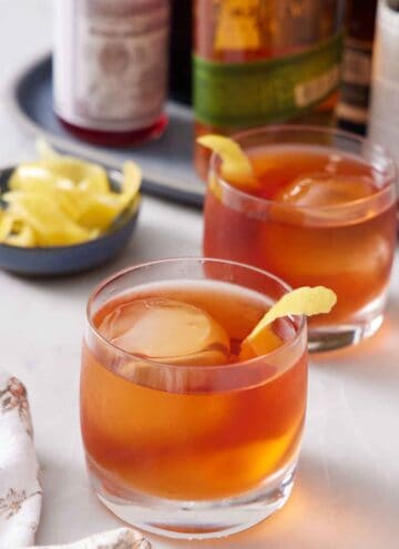 Two glasses of Vieux Carré cocktails with lemon peel garnish. A bowl of more lemon peels in the back along with bottles of alcohol.