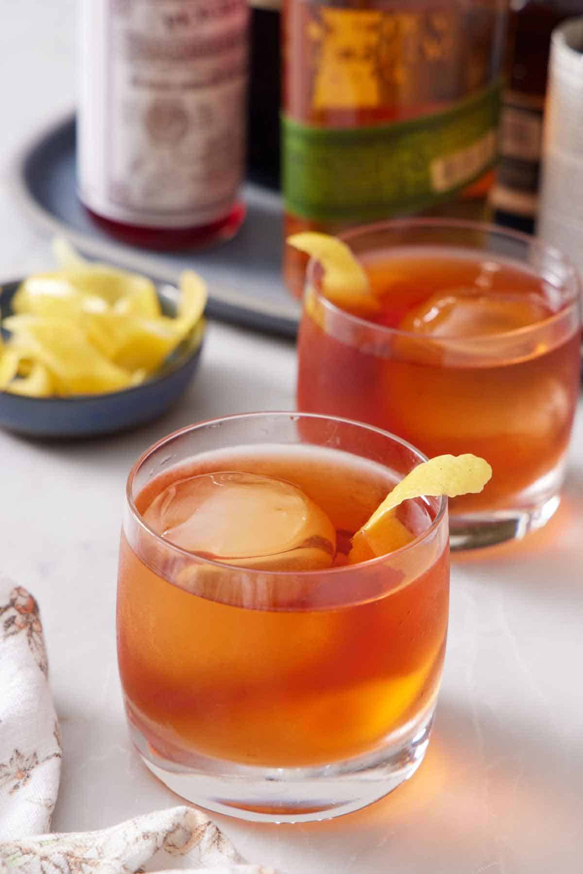 Two glasses of Vieux Carré cocktails with lemon peel garnish. A bowl of more lemon peels in the back along with bottles of alcohol.