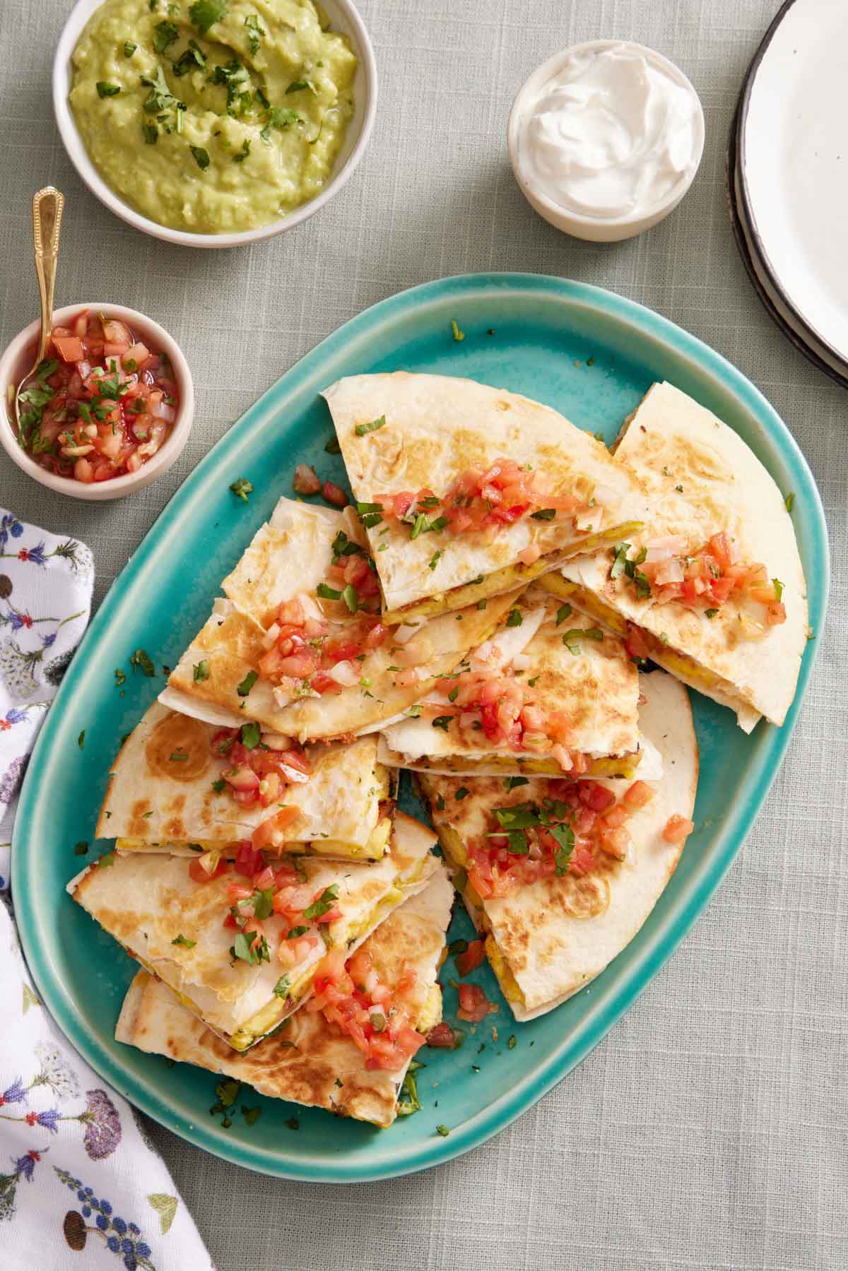 Overhead view of a platter of breakfast quesadillas topped with pico de gallo. A bowl of pico de gallo, guacamole, and sour cream on the side.