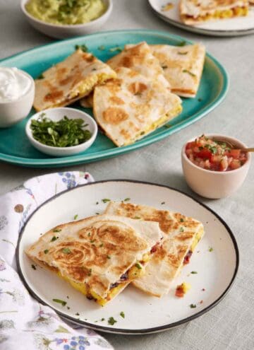 A plate with two pieces of breakfast quesadillas. A platter in the background with more with bowls of sour cream and chopped cilantro. A bowl of pico de gallo and guacamole in the background.