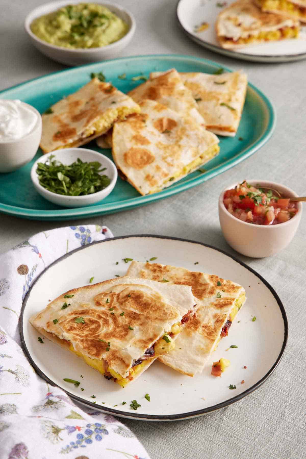 A plate with two pieces of breakfast quesadillas. A platter in the background with more with bowls of sour cream and chopped cilantro. A bowl of pico de gallo and guacamole in the background.