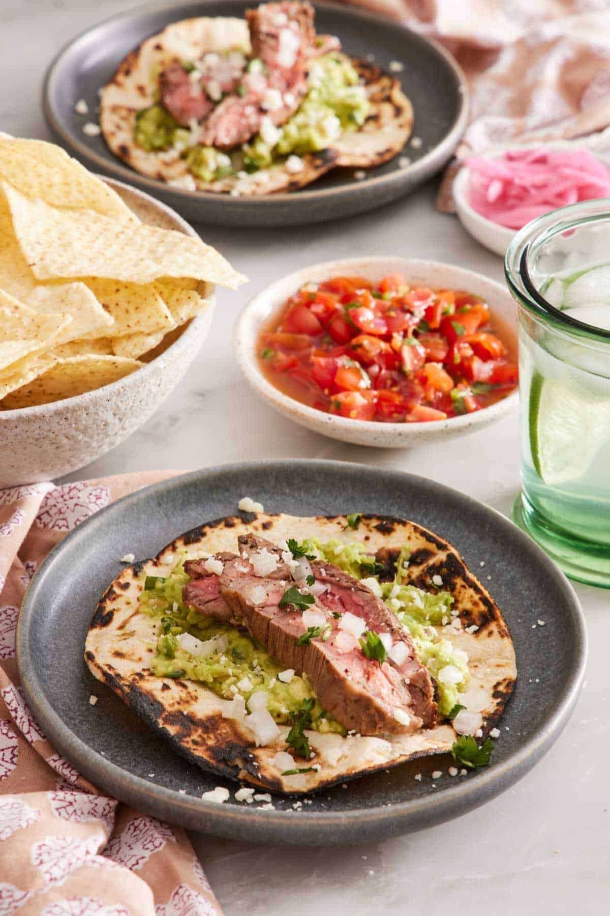 Carne asada taco on a plate with pico de gallo, chips, a drink, pickled onions, and another plated serving in the background.