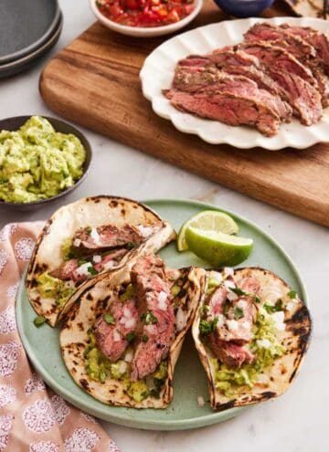 Three carne asada tacos on a plate with lime wedges. Guacamole, sliced grilled meat, pico de gallo, onions on a wooden serving board.