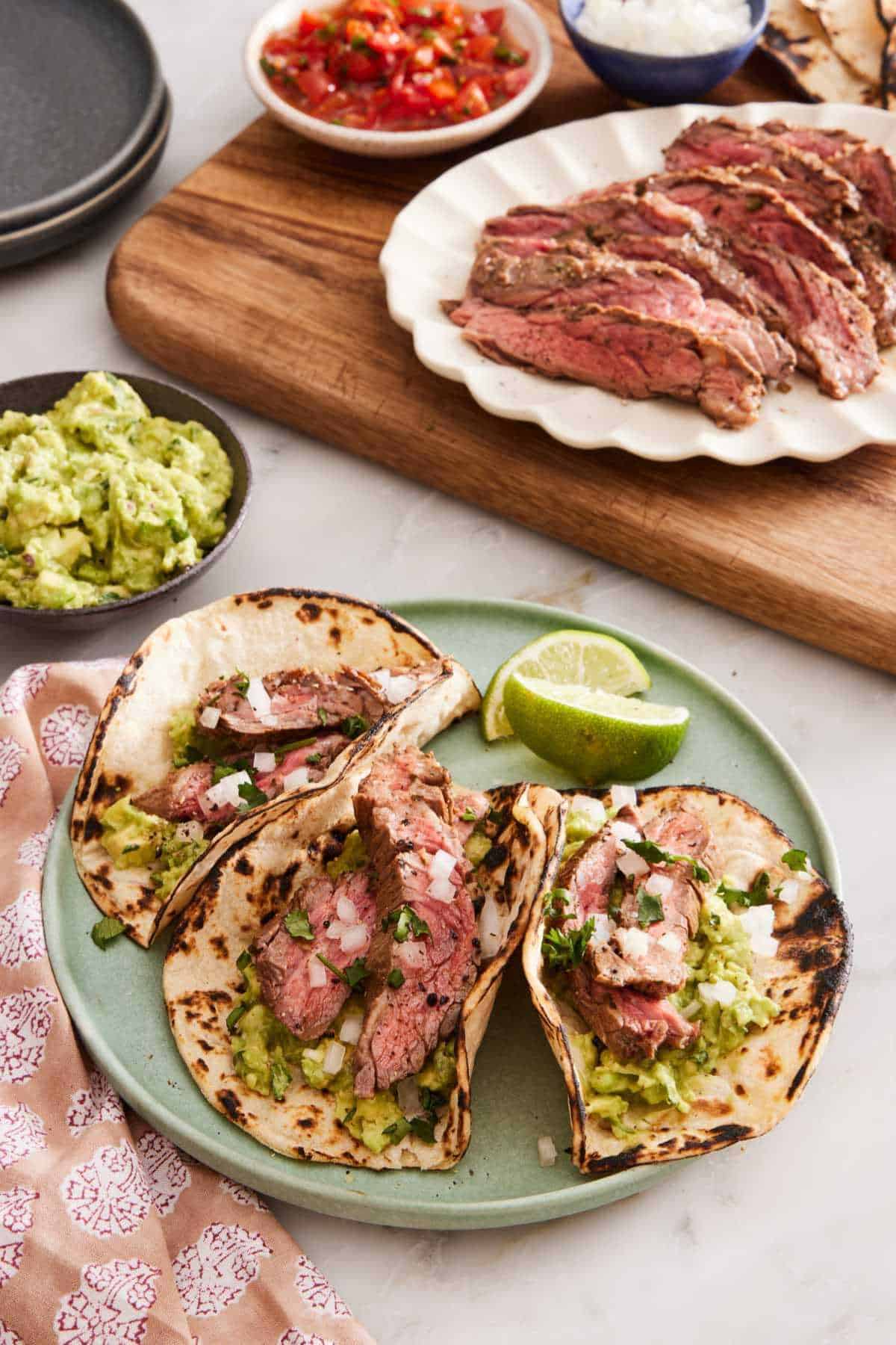 Three carne asada tacos on a plate with lime wedges. Guacamole, sliced grilled meat, pico de gallo, onions on a wooden serving board.