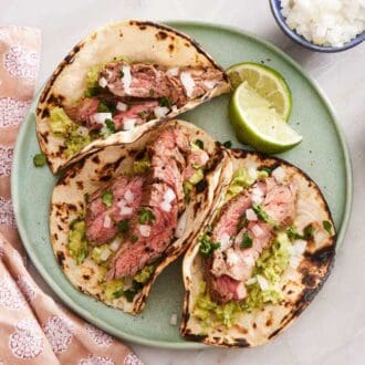 A plate with three carne asada tacos with lime wedges on the side.