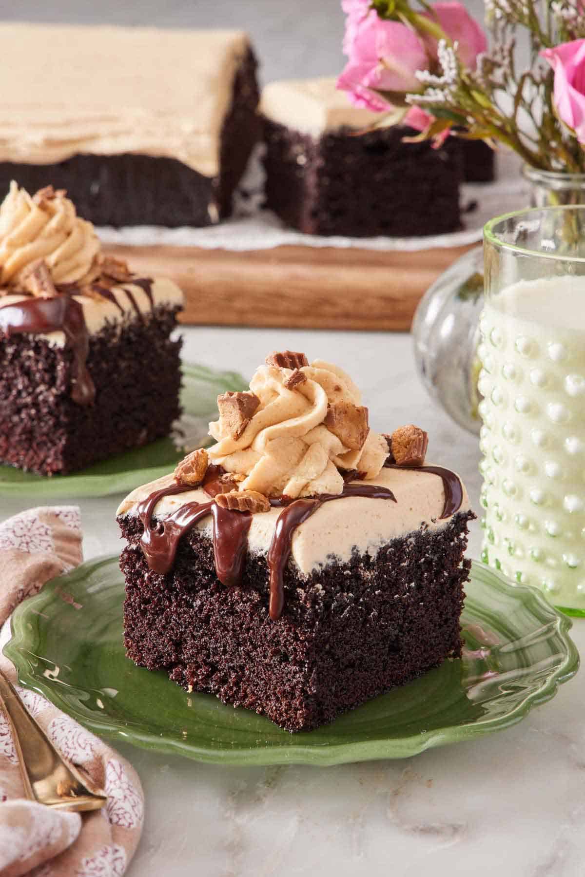 A square piece of chocolate peanut butter cake on a great plate. A class of milk, flowers, and the rest of the cake in the background.