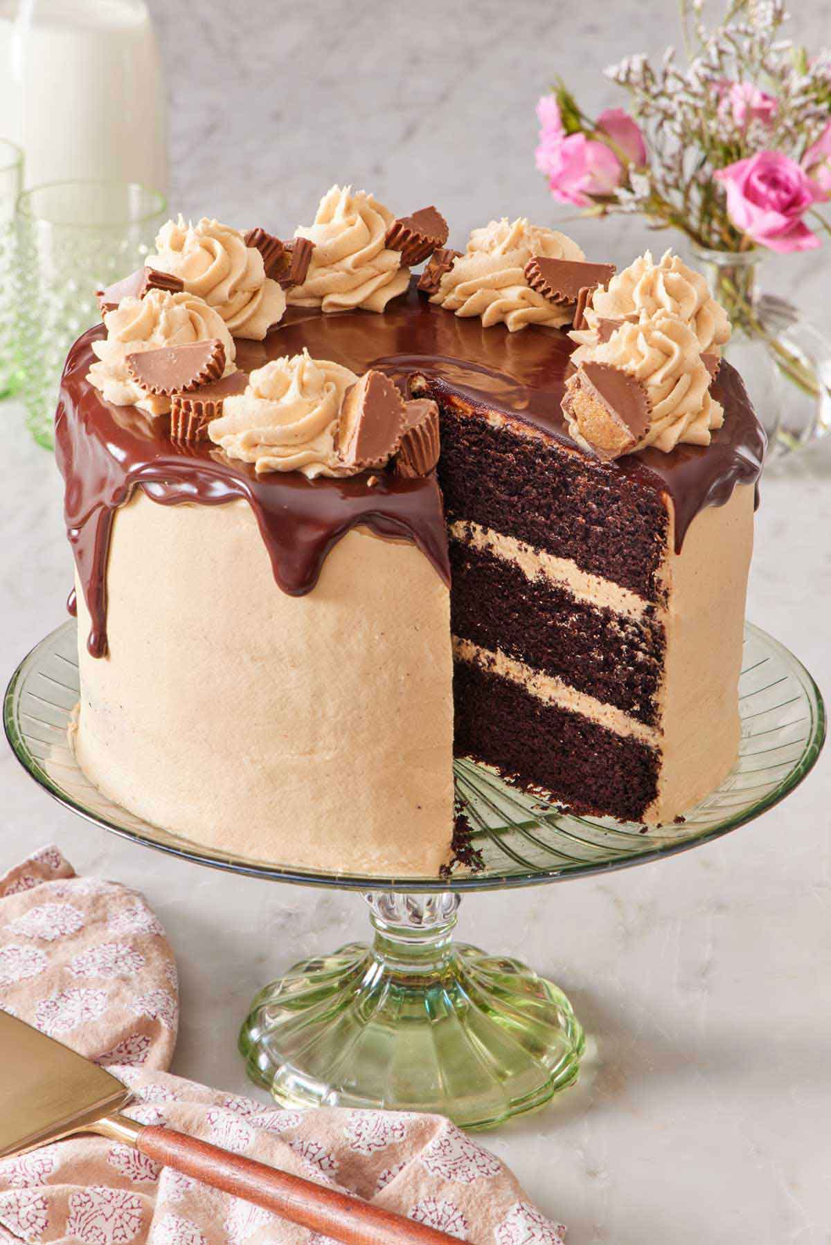 A chocolate peanut butter cake on a green cake stand with a slice cut out. Flowers in the background and a cake spatula in front.