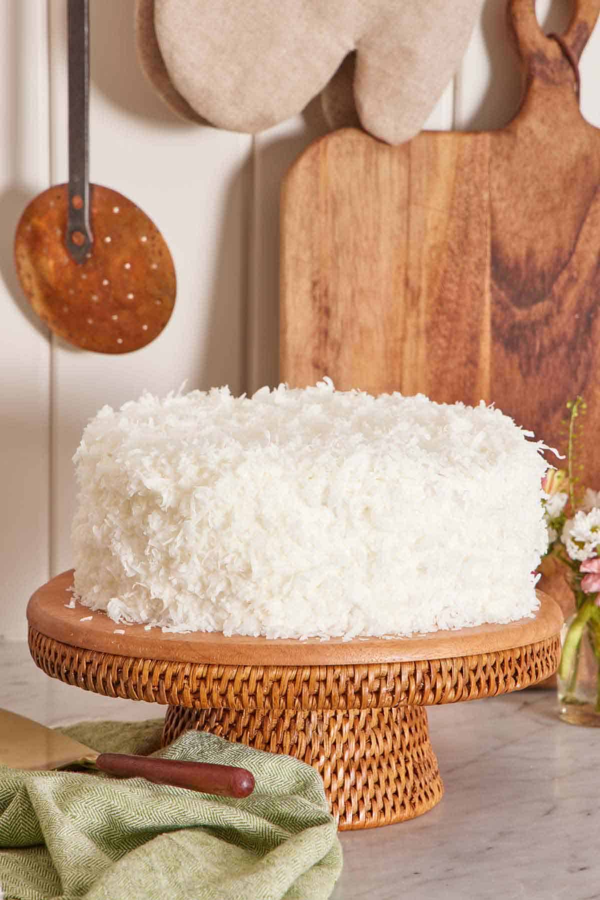 A coconut cake on a cake stand. A cake spatula on a linen napkin in front and a cutting board in the back.