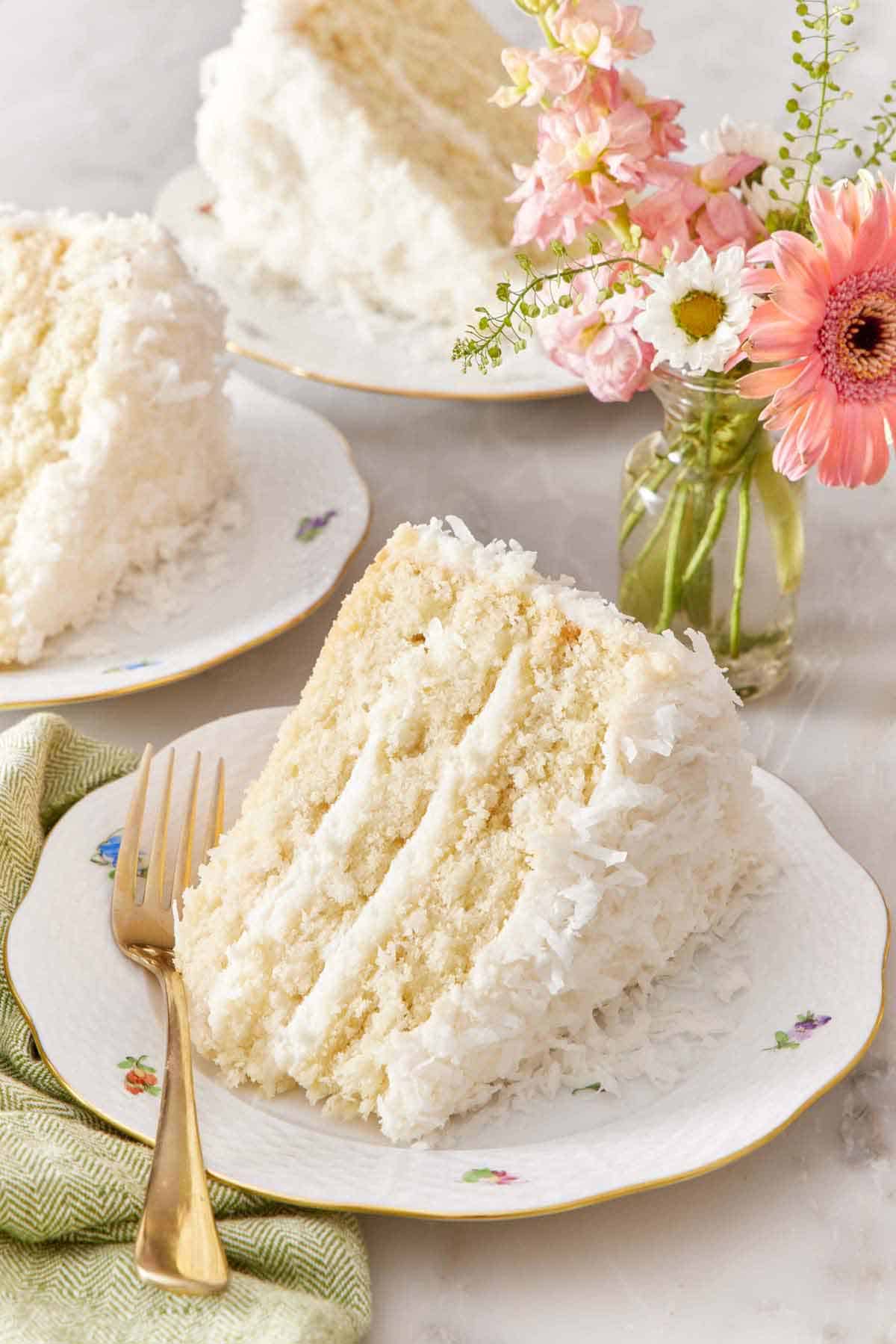A slice of coconut cake on its side on a plate showing the three layers with a fork beside it. Flowers in the background along with two slices of cake.