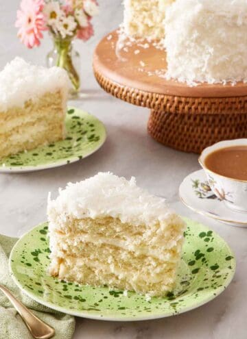 A slice of coconut cake on a plate. In the background is another plated slice and the rest of the cake on a cake stand.