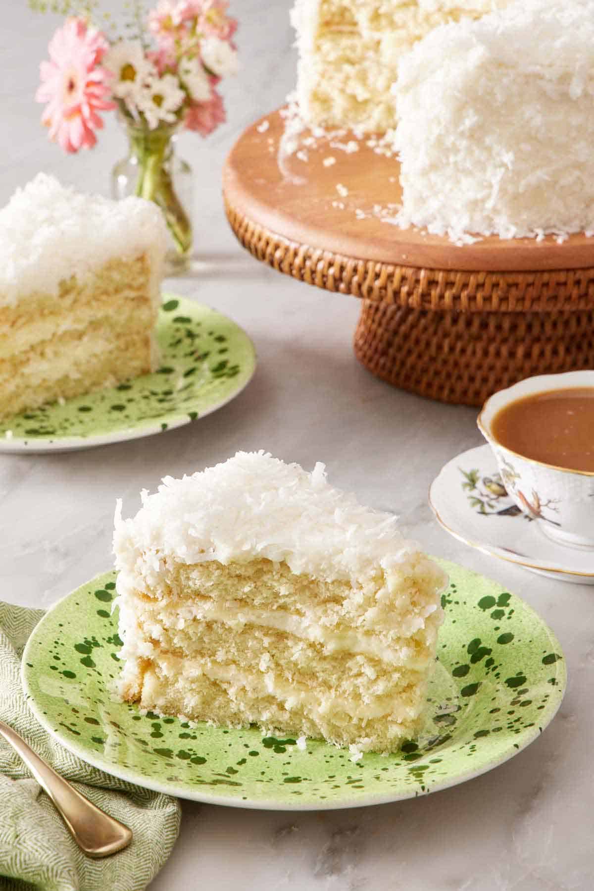 A slice of coconut cake on a plate. In the background is another plated slice and the rest of the cake on a cake stand.