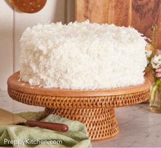 Pinterest graphic of a coconut cake on a cake stand. A cake spatula on a linen napkin in front and a cutting board in the back.