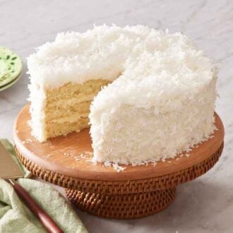 A coconut cake on a cake stand with a slice cut out. Flowers, plates, napkin, and a cake spatula on the side.