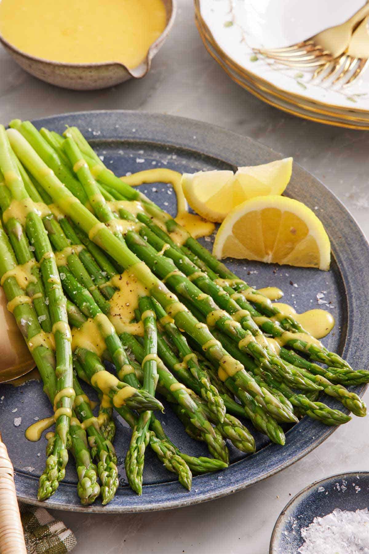 A platter of blanched asparagus with sauce drizzled on top and lemon wedges on the side. A bowl of sauce in the back along with some plates and forks.
