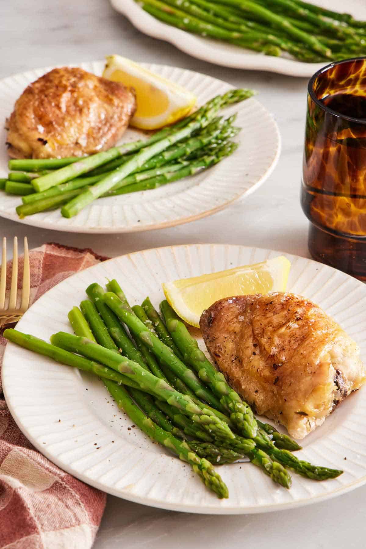 A plate with blanched asparagus, crispy chicken thighs, and a lemon wedge. Another plated serving in the background.