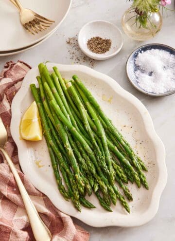 A platter of blanched asparagus with a lemon wedge. A bowl of salt, a bowl of pepper, vase of flower, plates, and forks on the side.