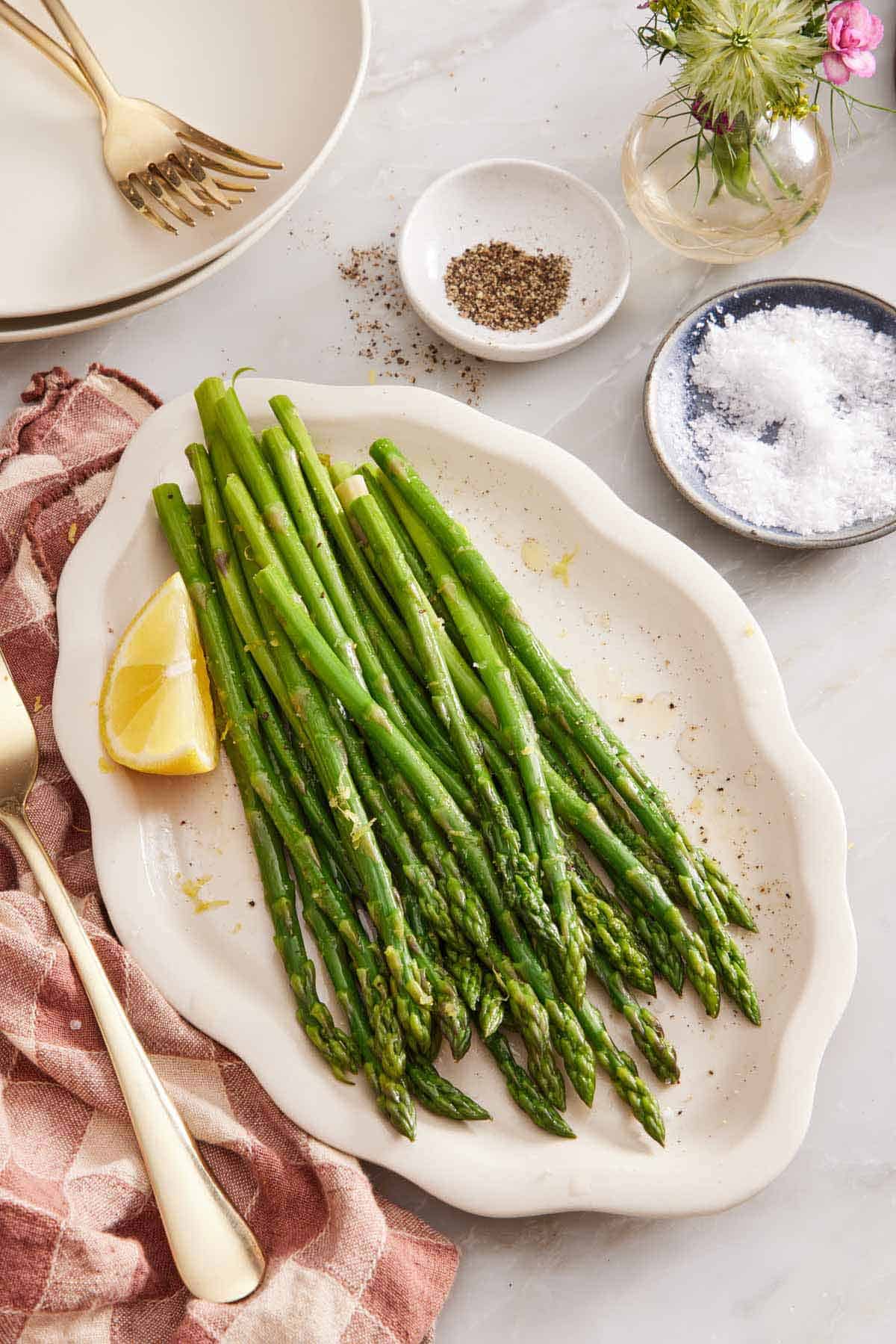 A platter of blanched asparagus with a lemon wedge. A bowl of salt, a bowl of pepper, vase of flower, plates, and forks on the side.