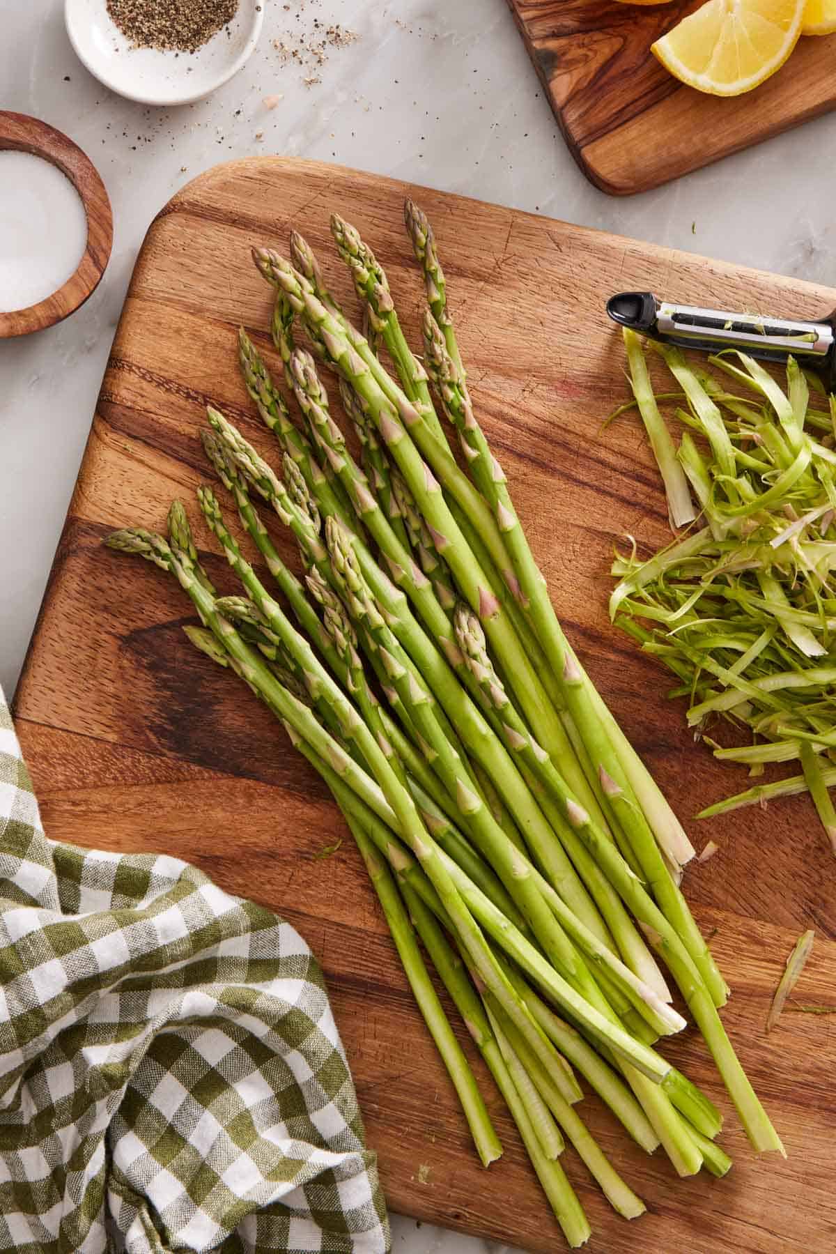 Asparagus with peeled ends on a wooden cutting board with the peeler and peeled bits on the side.