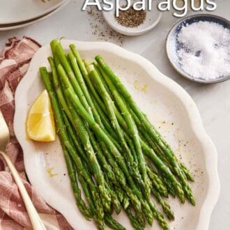 Pinterest graphic of blanched asparagus with a lemon wedge on a platter. A bowl of salt, a bowl of pepper, vase of flower, plates, and forks on the side.