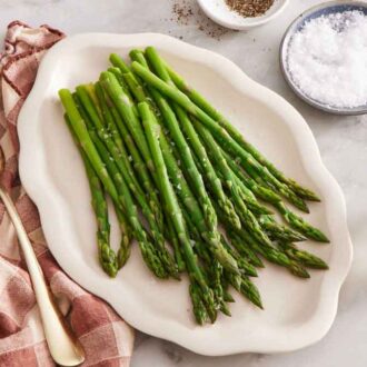 A platter of blanched asparagus with a vase of flower on the side along with a serving fork, salt, and pepper.