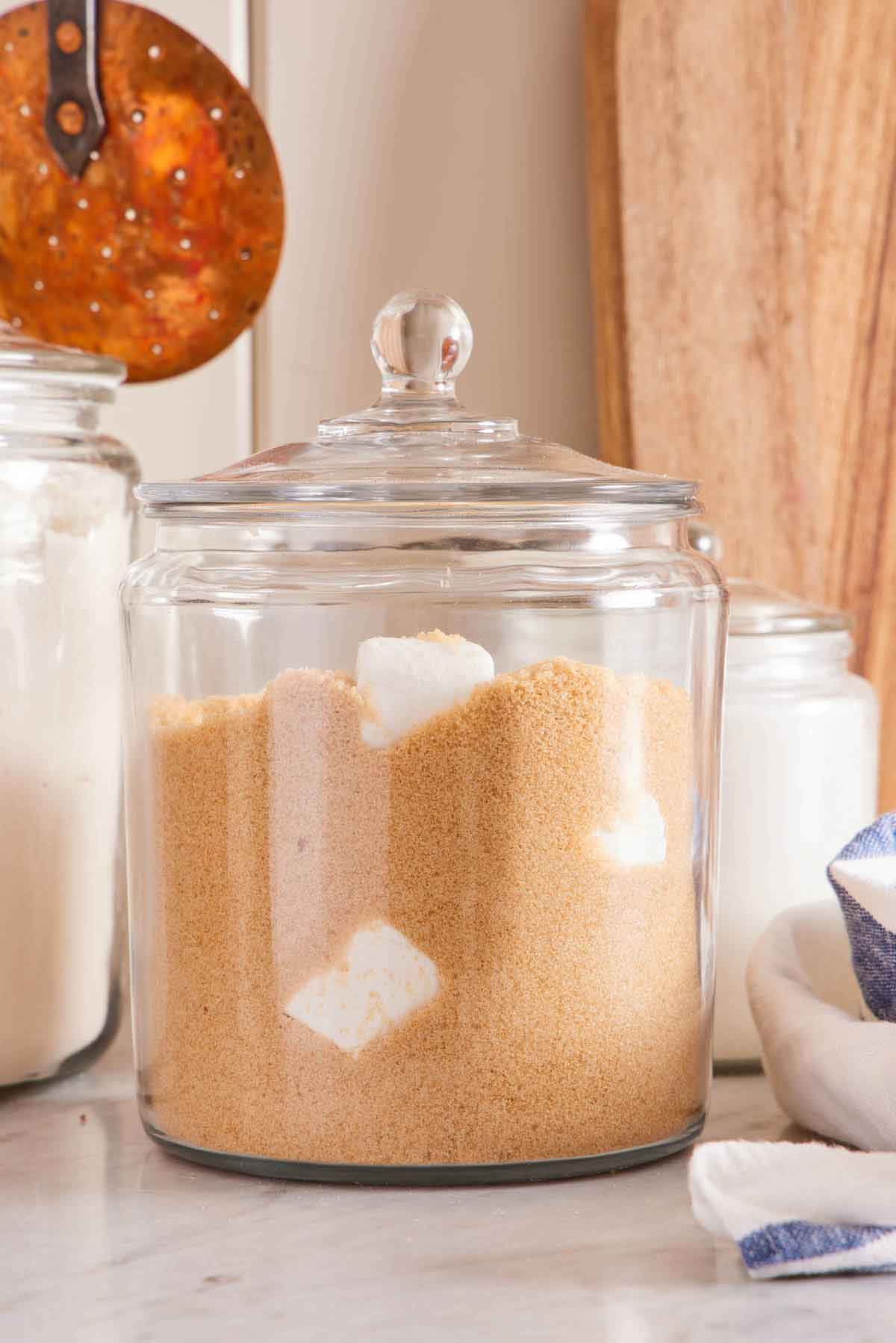 A large jar of softened brown sugar with saver tiles inside on a marble surface. More jars of kitchen staples in the back.