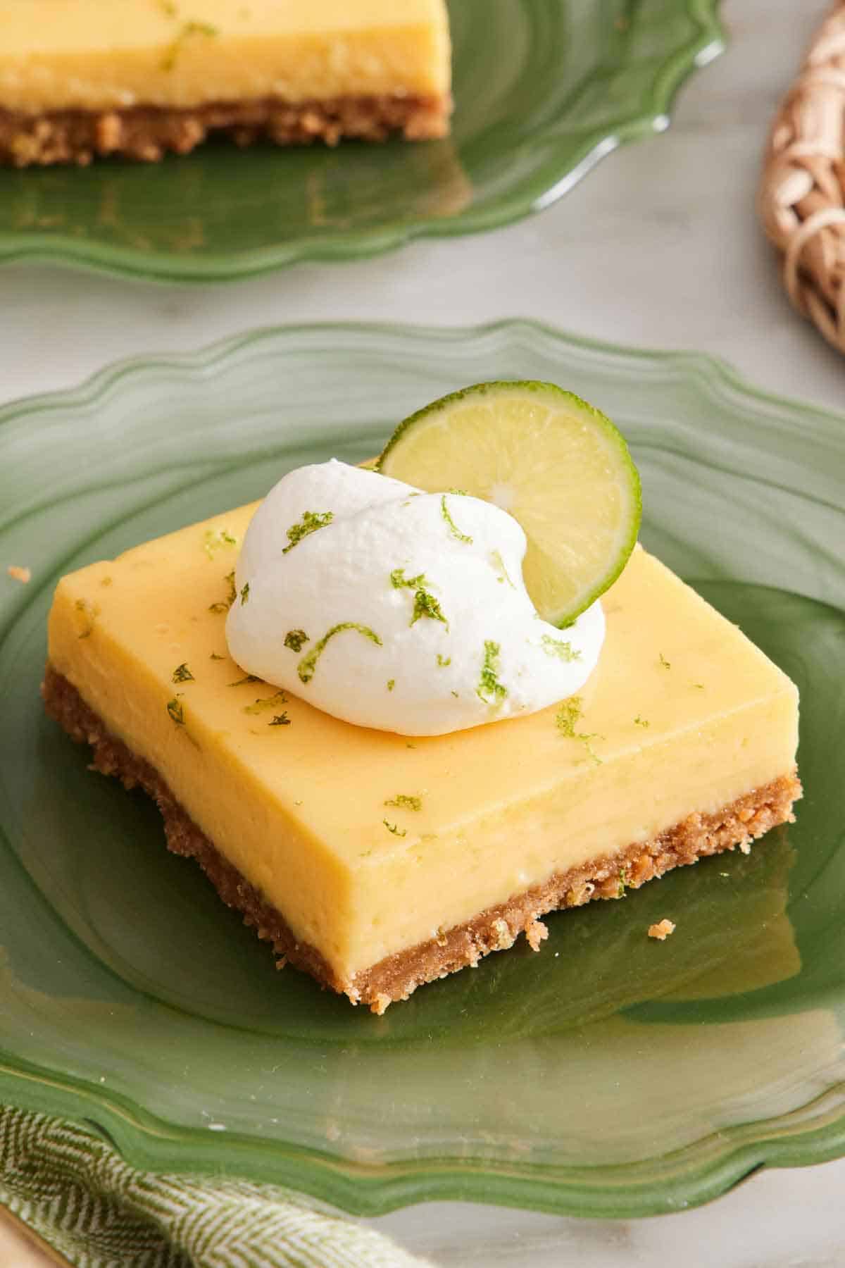 A piece of key lime pie bar on a green plate topped with whipped cream, lime zest, and a lime wheel.