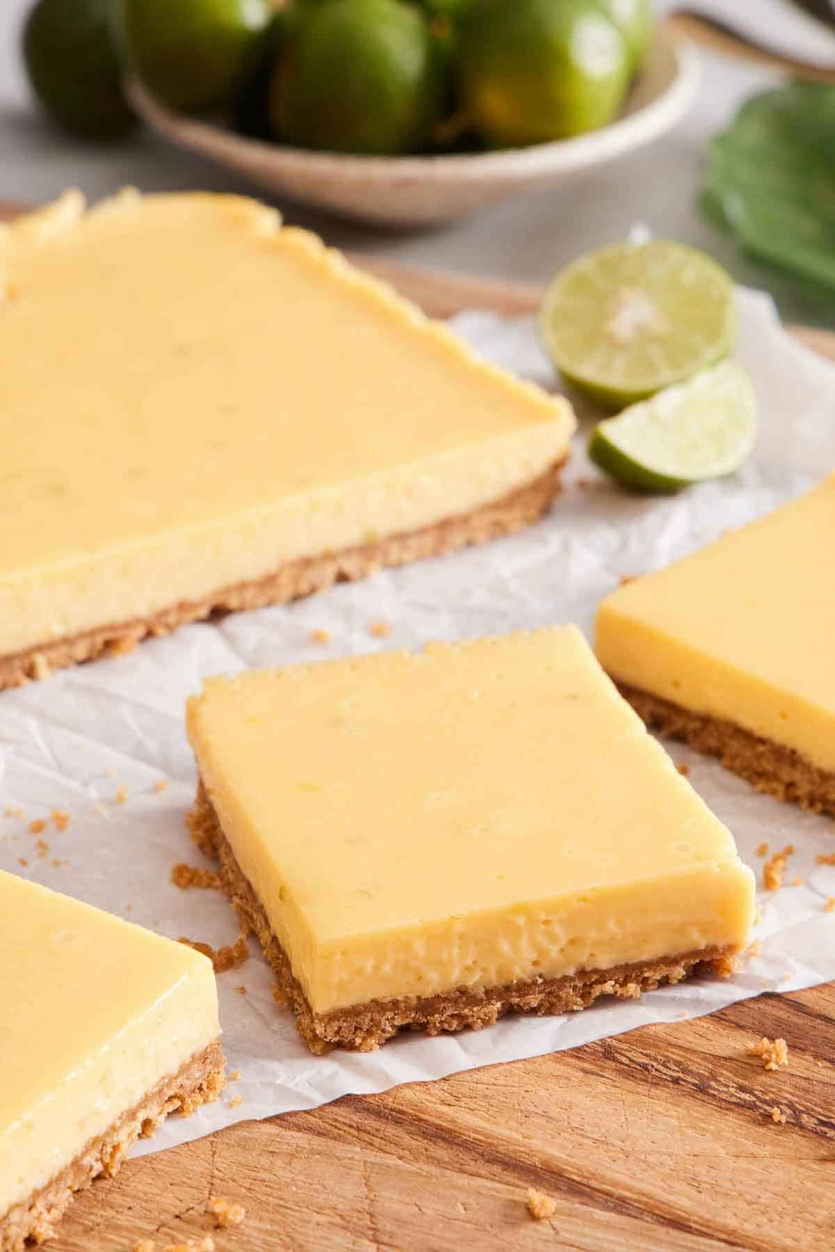 A few sliced key lime bars on a parchment lined board with the rest of the uncut dessert in the background along with some cut limes.