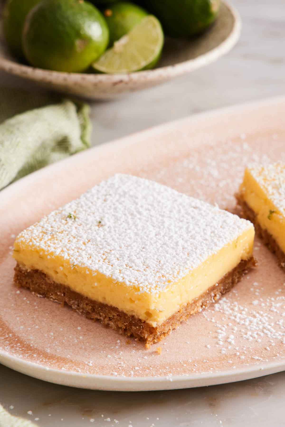 A key lime pie bar on a platter with a dusting of powdered sugar on top. Key limes on a plate in the background.
