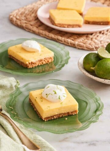 Two plates with key lime pie bars topped with whipped cream and lime zest. More slices in the background along with a plate of key limes.