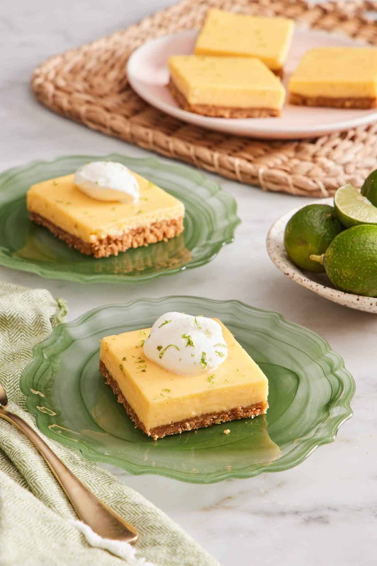 Two plates with key lime pie bars topped with whipped cream and lime zest. More slices in the background along with a plate of key limes.