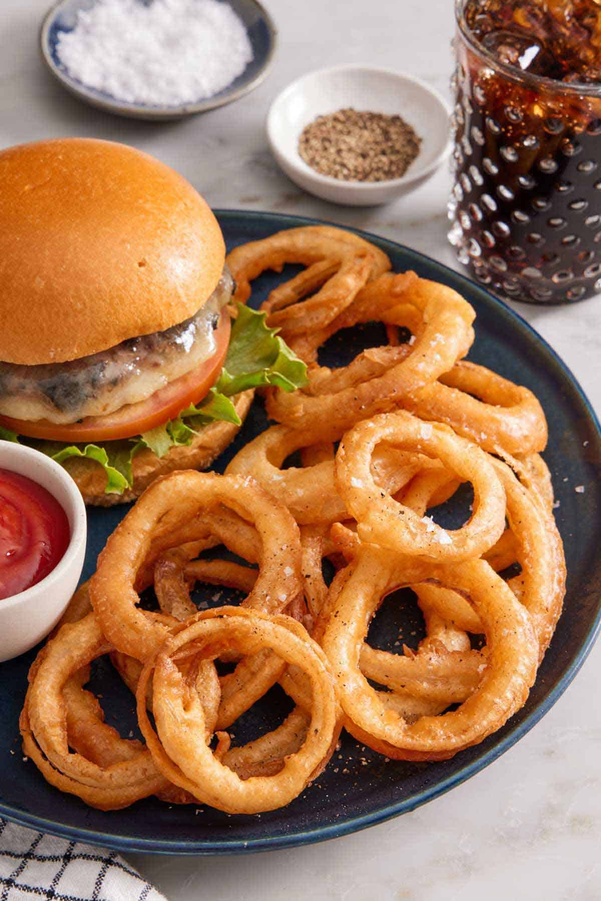 A plate of onion rings with a small bowl of ketchup and burger. A glass of soda, salt, and pepper in the back.