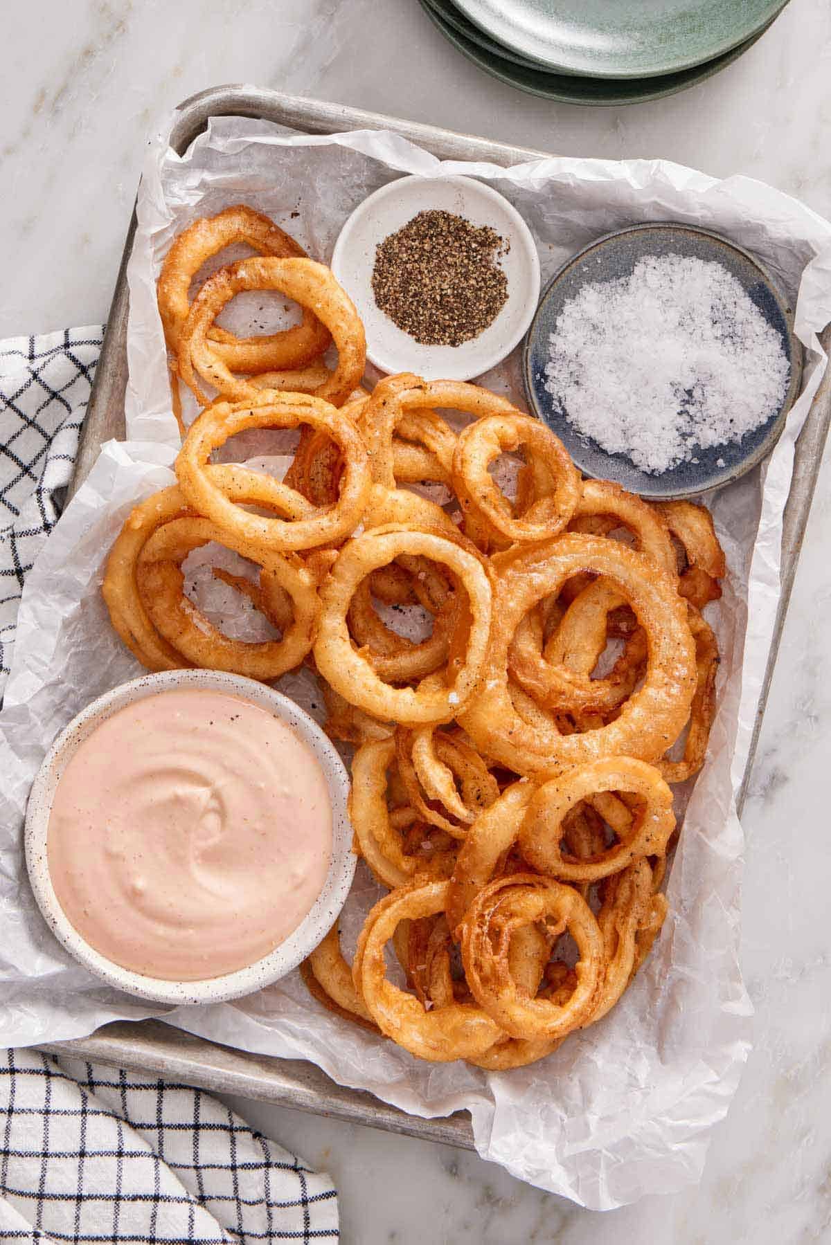 Overhead view of a lined baking sheet with onion rings, bowl of salt, pepper, and dipping sauce.