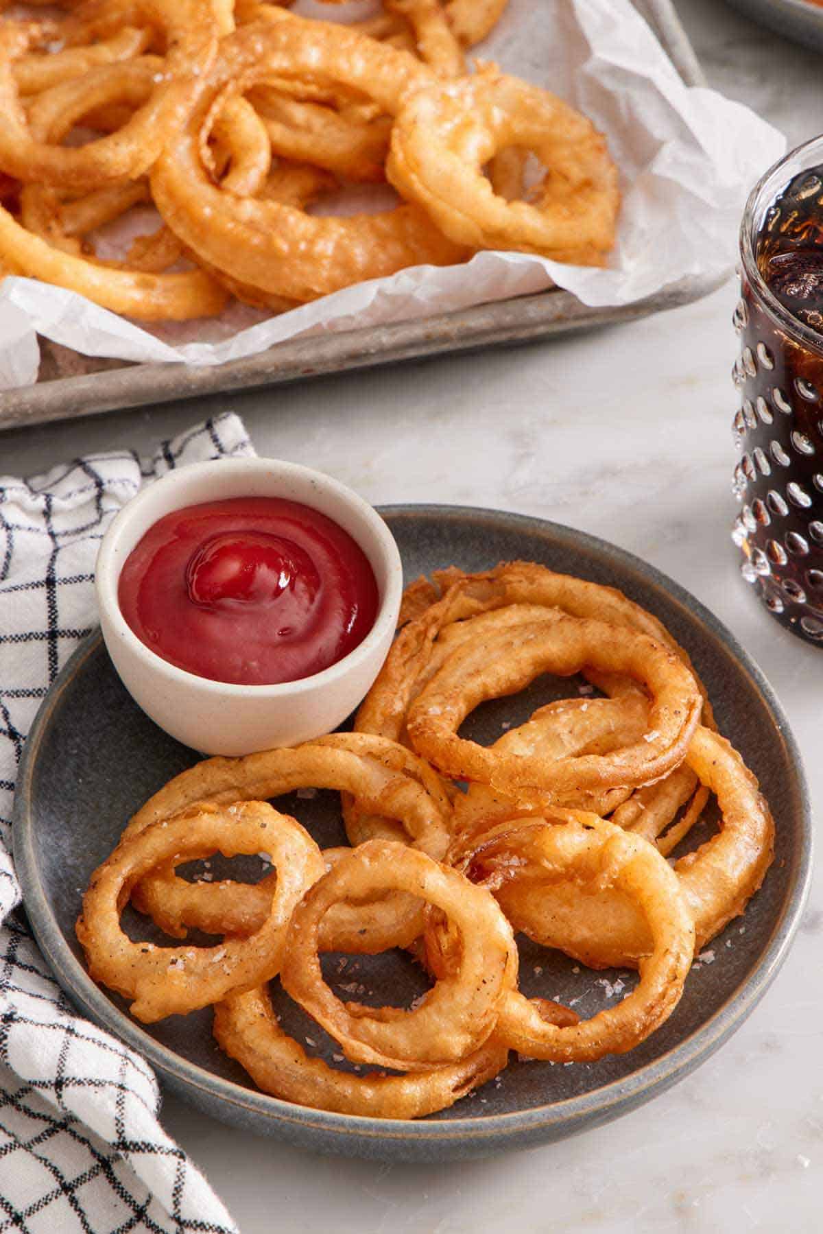 A plate of onion rings with onion rings with a bowl of ketchup. More onion rings in the background and a drink.