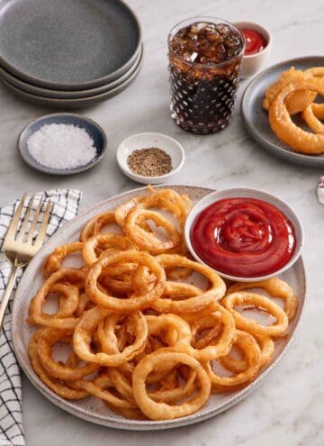 A platter of onion rings with a bowl of ketchup. A glass of soda, plate of onion rings, salt, pepper, and stack of plates in the back.