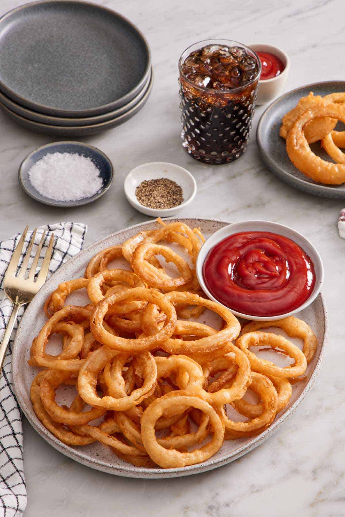 A platter of onion rings with a bowl of ketchup. A glass of soda, plate of onion rings, salt, pepper, and stack of plates in the back.