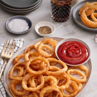 Pinterest graphic of a platter of onion rings with a bowl of ketchup. A glass of soda, plate of onion rings, salt, pepper, and stack of plates in the back.