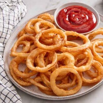 A platter of onion rings with a bowl of ketchup on the platter. A napkin on the side.